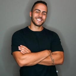 A smiling man with short dark hair and a trimmed beard, wearing a plain black crew-neck T-shirt, stands with his arms crossed against a solid light gray background; a small tattoo is visible on his left forearm, and he exudes confidence and approachability, fitting the context of a testimonial for Trainingym’s gym software as the founder of Action Black, a Colombian gym chain based in Spain.