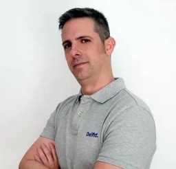 A professional head-and-shoulders portrait of José Manuel Delgado, owner of the personal training center Delfit in Granada, Spain, wearing a gray polo shirt with the blue 'Delfit' logo embroidered on the left chest, standing with arms crossed against a plain white background, looking directly at the camera with a confident, neutral expression, short dark hair with subtle graying at the temples, clean-shaven, and well-lit with soft studio lighting that highlights his facial features without harsh shadows.
