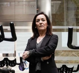 A confident woman with shoulder-length dark hair stands with her arms crossed in a modern fitness facility, wearing a black blazer over a patterned top, accessorized with a watch and bracelet, with gym equipment including black handles and a mask visible in the foreground, and a glass-paneled wall with horizontal blinds in the background, suggesting a professional and fitness-oriented environment.
